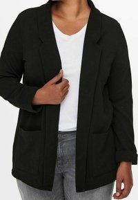 Musta rento leikkaus, pehmeästä materiaalista valmistettu bleiseri, jossa on käännetty kaulus, pitkät hihat ja kaksi etutaskua. Päällä valkoinen toppi.