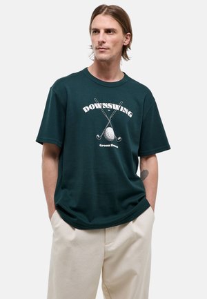 Jeune homme aux cheveux mi-longs portant un t-shirt vert foncé avec des clubs de golf croisés et le texte « Downswing at Green Home ».