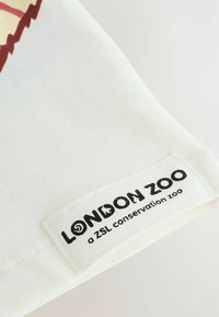 Tela de algodón blanco con una textura suave. Presenta una etiqueta tejida en texto negro: "LONDON ZOO un zoológico de conservación de ZSL" en el borde inferior.