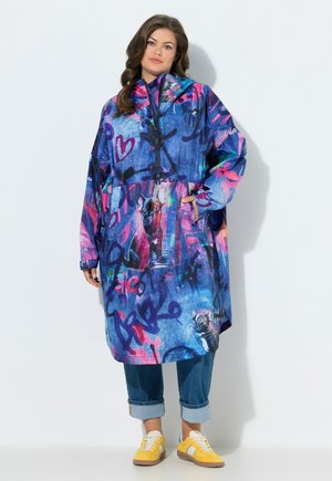 Nainen, jolla on värikäs, ylipitkä huppari, jossa on graffiteja ja koirakuvioita, käännetyt farkut ja keltaiset tennarit, seisoo valkoista taustaa vasten.