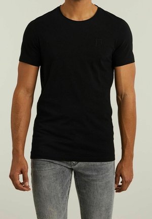 Homme portant un t-shirt noir uni à manches courtes et un jean gris clair, debout devant un fond neutre.