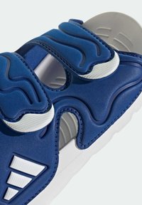 Granatowy trampek z teksturowanym materiałem i białymi akcentami. Charakteryzuje się unikalnym falistym wzorem, podwójnymi paskami na rzep oraz gumową podeszwą.