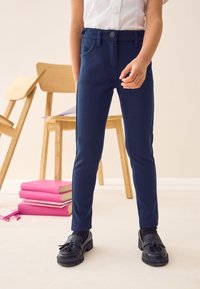 Pantaloni blu navy realizzati in tessuto elastico, con chiusura a bottone, tasche laterali e vestibilità slim, indossati con mocassini neri e calzini scuri.