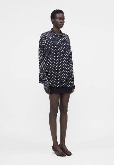 Ψηλή γυναίκα με κοντά μαλλιά φοράει ένα φαρδύ μαύρο πουκάμισο με λευκά σχέδια, μια μαύρη μίνι φούστα και μαύρα σανδάλια με λουράκι στο πίσω μέρος.