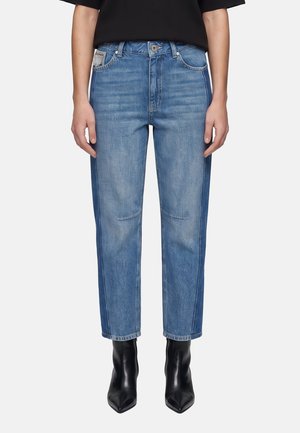 Person trägt blaue Jeans mit geradem Bein und Seitenteilen sowie schwarze spitze Stiefeletten, die Hände ruhen seitlich.