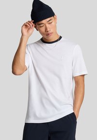 Jonge man in een wit t-shirt met een klein embleem, marineblauwe broek, een marineblauwe gebreide muts en een zilveren ketting, poseert tegen een grijze achtergrond.