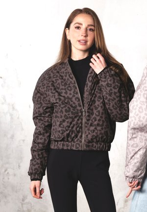 Young woman wearing a dark gray leopard print zip-up jacket with elastic cuffs and black pants, standing against a light textured background.