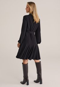 Donna con lunghi capelli biondi, che indossa un vestito nero a maniche lunghe con pieghe e stivali scuri fino al ginocchio, in piedi davanti a uno sfondo neutro.