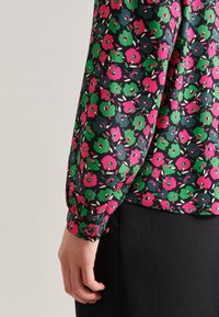 Blusa a maniche lunghe in tessuto nero con vivaci motivi floreali nei colori rosa, verde e bianco, con una texture liscia e polsini attillati.