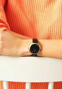 Schwarze Analog-Uhr mit goldenen Akzenten und braunem Lederarmband, präsentiert auf einem hellhäutigen Arm vor einem Hintergrund aus einem orangefarbenen strukturierten Pullover.