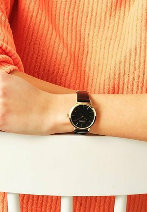 Schwarze Analog-Uhr mit goldenen Akzenten und braunem Lederarmband, präsentiert auf einem hellhäutigen Arm vor einem Hintergrund aus einem orangefarbenen strukturierten Pullover.