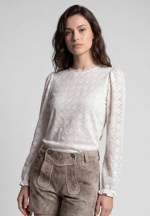 Vrouw met lang donker haar die een witte kanten top met lange mouwen en beige suède shorts met bloemenborduurwerk draagt, staand tegen een effen achtergrond.