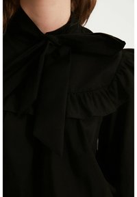 Blusa nera con colletto ampio e fiocco, dettagli arricciati lungo il scollo e texture del tessuto morbida e liscia.