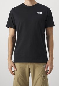 Muž v černém tričku s krátkým rukávem The North Face a béžových cargo kalhotách se zipovými kapsami, stojící na neutrálním pozadí.