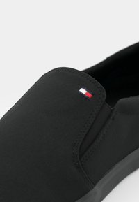 Schwarzer Schlupfschuh mit glattem Textilobermaterial, elastischen Seitenteilen und kleinem dreifarbigen Logo an der Seite. Strukturierte schwarze Sohle.