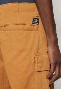Cargo-Hosen in einem sandfarbenen Farbton mit glatter Textur. Zu den Merkmalen gehören eine seitliche Tasche, eine Gesäßtasche und ein schwarzes Timberland-Logoetikett.