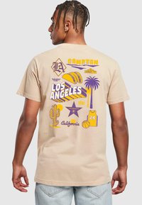 Camiseta de algodón beige con estampado gráfico multicolor en la parte posterior, que incluye diseños de "Los Ángeles", palmera, baloncesto y taco.