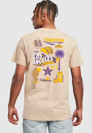 Camiseta de algodón beige con estampado gráfico multicolor en la parte posterior, que incluye diseños de "Los Ángeles", palmera, baloncesto y taco.