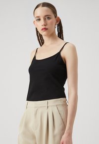 Black sleeveless tank top made of soft fabric with thin straps, paired with high-waisted beige pleated trousers. Clean lines and minimal design.