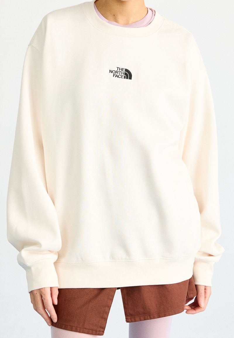 Personne portant un sweat-shirt ample de couleur blanc cassé avec un petit logo noir The North Face sur la poitrine et une jupe marron avec une fente à l'avant.