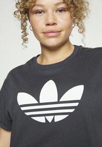 Juodas medvilnės t-shirt su dideliu baltu Adidas logotipu ir trimis juostelėmis po juo. Audinys turi lygų, tekstūrinį paviršių.