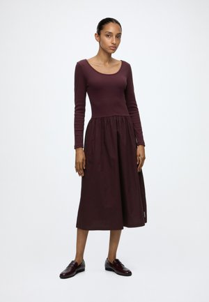 Woman standing in a long-sleeve, burgundy midi dress with a gathered skirt and matching dark burgundy loafers on a white background.