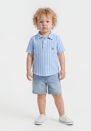 Νήπιο με ξανθά κατσαρά μαλλιά, που φορά μπλε και λευκή ριγέ μπλούζα με γιακά, τζιν σορτς και άσπρα αθλητικά παπούτσια, στέκεται σε λευκό φόντο.