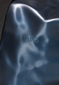 Materiale blu scuro e liscio con la parola "RAINS" in rilievo, che indica il marchio. La texture sembra lucida e leggermente riflettente.