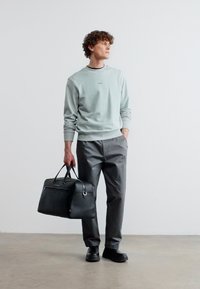 Νέος άνδρας με καθημερινή στολή που κρατά μια μαύρη δερμάτινη τσάντα, στέκεται σε τσιμεντένιο δάπεδο μπροστά σε έναν απλό λευκό τοίχο, κοιτώντας προς τα πλάγια.