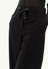 Czarne spodnie o dopasowanym kroju z gładką teksturą, z detalem w postaci kokardy w talii, bocznymi kieszeniami oraz dopasowanym fasonem.