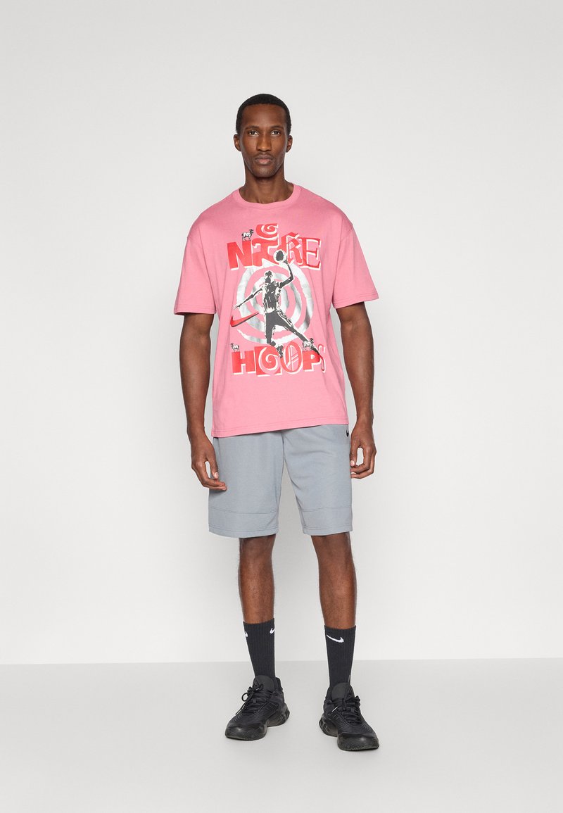 T-shirt di cotone rosa con stampa grafica, abbinata a pantaloni corti grigi. Scarpe da ginnastica nere e calze con dettaglio del logo bianco completano l'outfit.