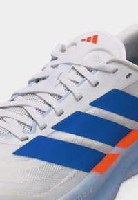 Baskets blanches avec des bandes Adidas bleues, des lacets blancs, des accents orange sur la semelle intermédiaire et un détail en mesh respirant sur la tige.