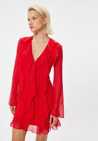 Rotes Chiffonkleid mit Rüschendetails, tiefem V-Ausschnitt und langen Ärmeln. Der Stoff weist kleine gepunktete Muster auf und hat einen fließenden Rock.
