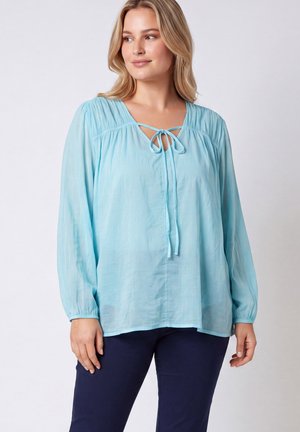 Vrouw draagt een losse lichtblauwe blouse met lange mouwen en een strikdetail vooraan en donkerblauwe broek tegen een effen achtergrond.