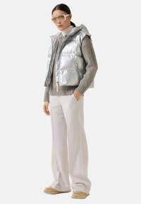 Vrouw in oversized beige broek, grijze gebreide trui, zilveren puffer vest, tan instappers, en grote beige getinte zonnebril.