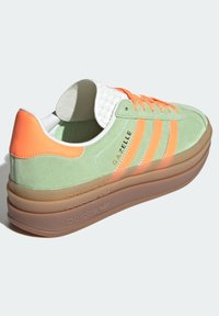 Zelené semišové tenisky s oranžovými akcenty, bílým jazykem a třemi oranžovými pruhy. Mají silnou průhlednou podrážku s brandingem Adidas.