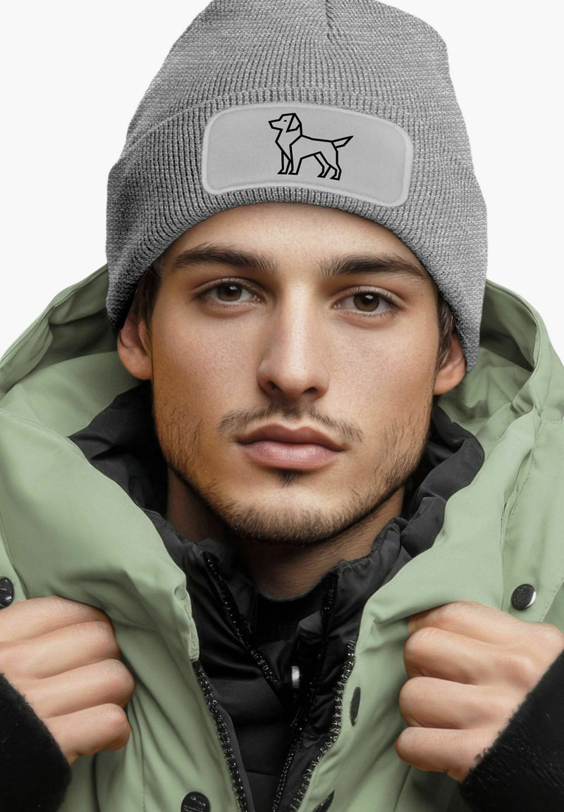 Gray knitted beanie with a flat panel featuring a black outlined dog design. Worn with a green puffer jacket, showcasing textured materials.