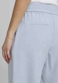 Boční pohled na osobu v světle modrých elastických kalhotách s rozparkem na zadní kapse, bílé tričko částečně viditelné, ruka opřená podél těla.