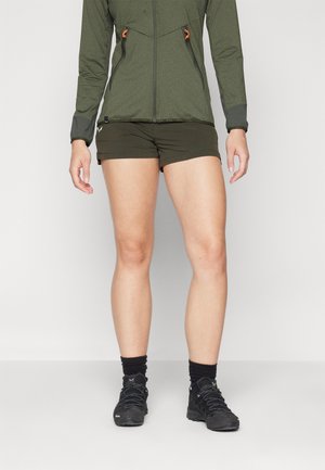 Woman wearing olive green zip-up jacket, dark green shorts, black socks, and black hiking shoes standing on a white floor.