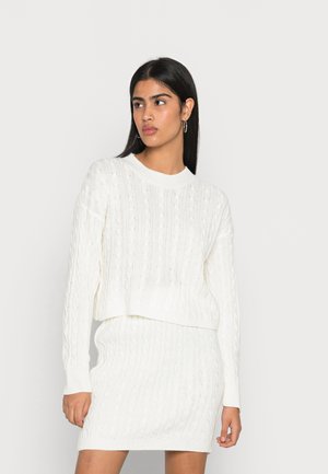 Suéter blanco de punto con un diseño corto y mangas largas, combinado con una falda de punto trenzado a juego, que presenta patrones texturizados en toda su extensión.