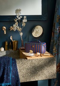 Blue and orange striped cosmetic bag on wooden surface, surrounded by gold accents, glass vase, round mirror, and textured dark towel.