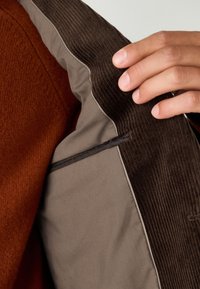 Doublure de veste en velours côtelé marron avec un tissu beige lisse. Présente un motif à carreaux bleu décoratif le long du bord d'une poche cachée.