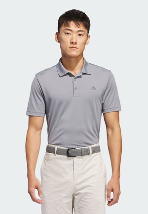 Homme portant un polo Adidas gris clair rentré dans un pantalon blanc avec une ceinture tissée grise, se tenant devant un fond uni.
