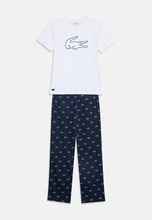 Chemise blanche à manches courtes avec grand logo crocodile, associée à un pantalon bleu marine arborant un motif répétitif du logo crocodile.