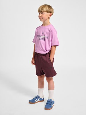 Ung dreng stående med hænderne i lommerne iført en lilla 'Serve the Game' T-shirt, vinrøde shorts, hvide sokker og blå sneakers.