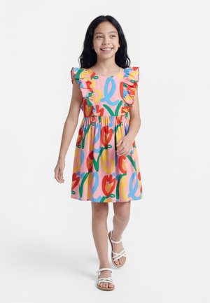 Dekle hodi, oblečeno v pisano obleko brez rokavov z naborki in belimi sandali, se smehlja na enobarvnem belem ozadju.