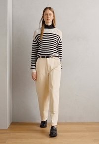 Stripet strikkegenser i svart og hvitt, kremfargede bukser og svarte sko. Modellen står foran en nøytral bakgrunn med tregulv.