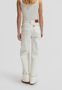 Femme portant un jean blanc taille haute à jambes larges avec des revers, un haut beige sans manches, debout devant un fond uni.