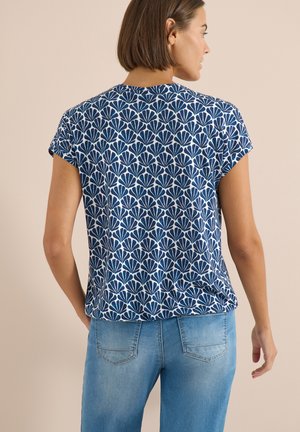 Femme vue de dos portant un haut à manches courtes à motifs bleus et blancs et un jean en denim bleu clair, sur un fond beige uni.