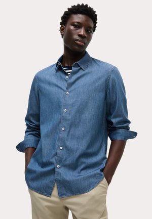 Camisa de mezclilla azul con botones y mangas largas, con cuello y dobladillo recto; llevada sobre una camisa de rayas; textura suave, ajuste casual.